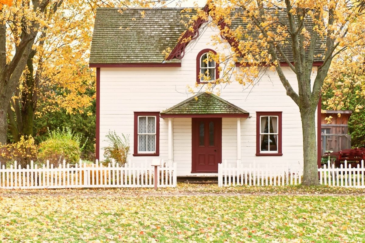 a warm, homey, white and red house in the fall with a white fence, grass and trees around it