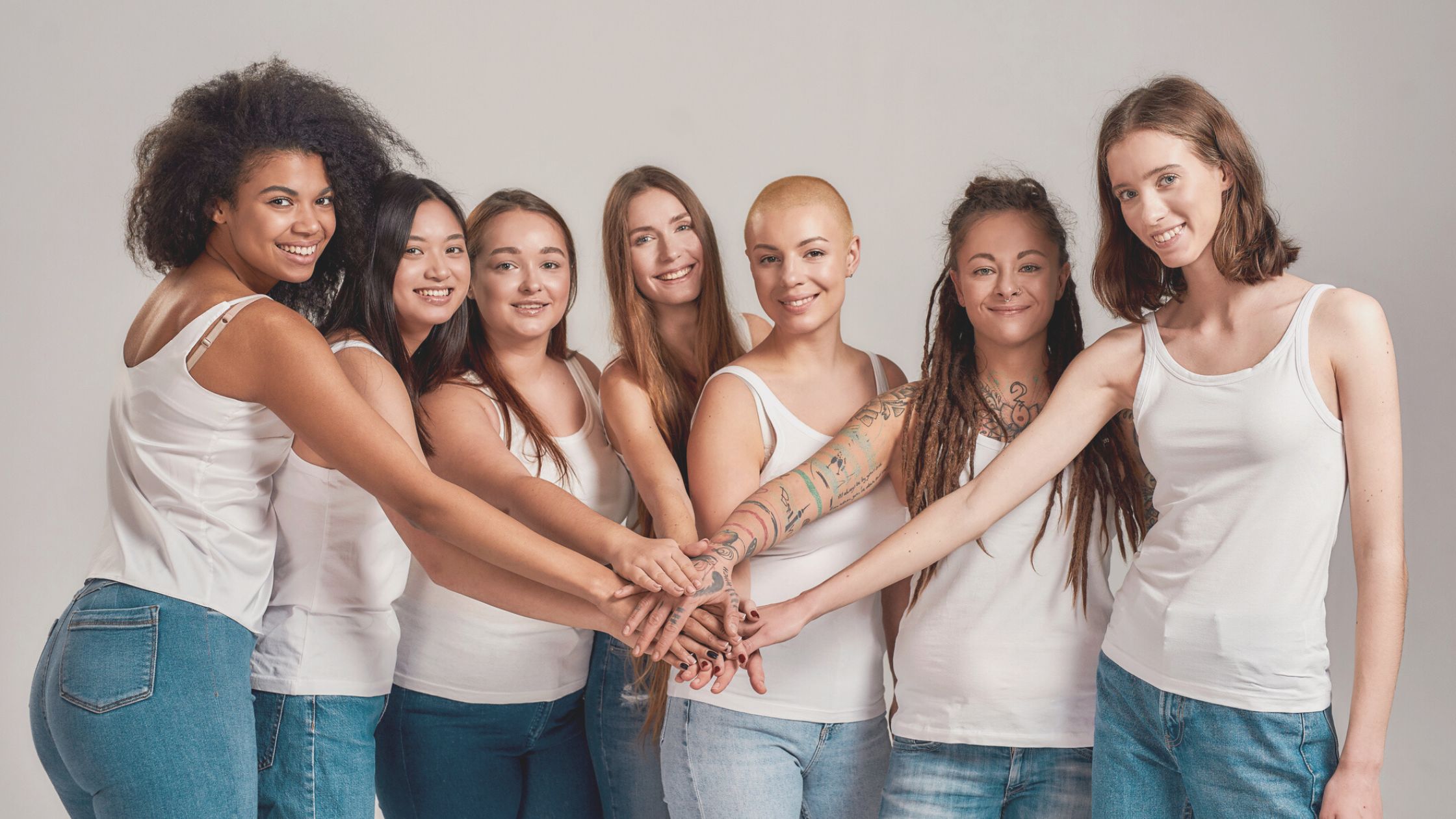 diverse group of women in white tank tops and denim jeans in a line, putting their hands together