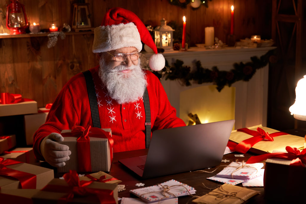 Happy old Santa Claus wearing hat holding gift box recording a video on his laptop computer sitting at workshop home table late on Merry Christmas eve.