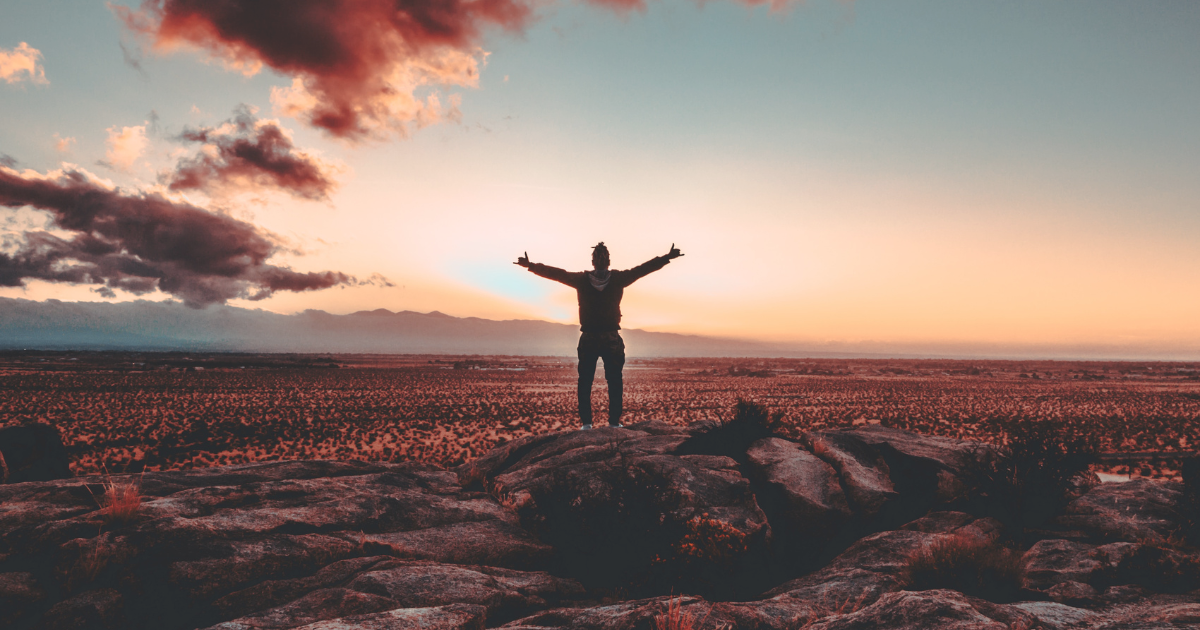 Person standing on a boulder in the desert with their arms raised and a beautiful sunset in the background